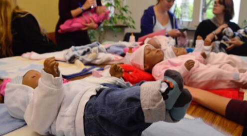 Tisch auf dem Baby-Simulationspuppen liegen. Im Hintergrund Schülerinnen, die jeweils eine Puppe im Arm halten und ein Fläschchen geben.