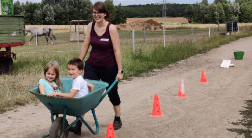 Feldweg an einer Pferdeweide, Pylonen markieren einen Parcour, den eine junge Frau mit Schubkarre fährt, in der Schubkarre sitzen ein Mädchen und ein Junge.