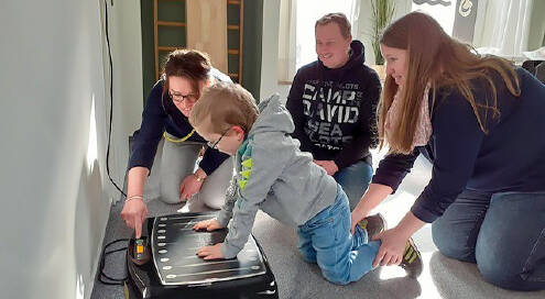 Junge kniet auf dem Boden, Hände auf einem Therapiegerät. Mutter hält den Jungen an den Beinen fest, Vater sitzt im Hintergrund, Janina Brunkhorst drückt auf einen Knopf am Therapiegerät.