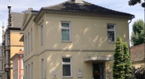 Waisenhaus Gebäude mit hellgelber Fassade, Büsche im Vordergrund, blauer Himmel im Hintergrund.