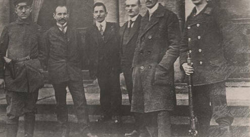 Schwarz-weiß Foto einer Gruppe von sechs Männern. Es handelt sich um den Arbeiter- und Soldatenrat. Sie tragen dunkle Mäntel und Stiefel, einer hat ein Gewehr vor sich abgestellt.