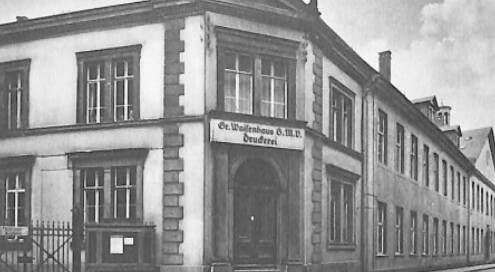 Schwarz-weiß Foto der Waisenhaus Druckerei. Man sieht Eingang, Hausfassade, Fenster und einen Zaun.