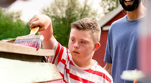 Junge mit Down-Syndrom streicht Holzhaus mit roter Farbe an. Ein Mann steht lachend hinter ihm, eine Frau schaut links um die Ecke. Im Hintergrund stehen Bäume.
