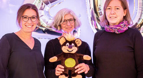 Drei Frauen in schwarzer Kleidung (links Janina Brunkhorst, rechts Sandra Hüsemann), Frau in Mitte hält braunes Wolli-Kuscheltier mit grünem Bauch und lila Nase, Hintergrund silberne Luftballons in Form einer 20.