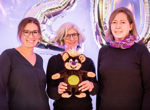 Drei Frauen in schwarzer Kleidung (links Janina Brunkhorst, rechts Sandra Hüsemann), Frau in Mitte hält braunes Wolli-Kuscheltier mit grünem Bauch und lila Nase, Hintergrund silberne Luftballons in Form einer 20.