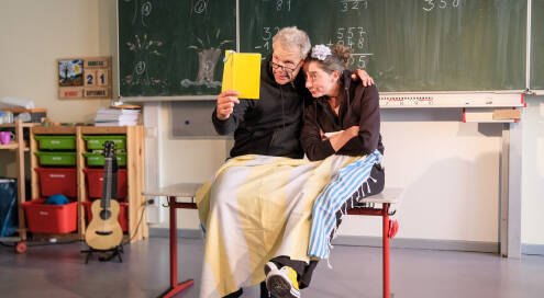 Zwei Schauspieler, links Mann, rechts Frau, sitzen zugedeckt im Klassenraum auf Tisch. Mann hält ein Buch, beide gucken hinein, Mann hat Arm um Frau gelegt, Frau hält Arme verschränkt.