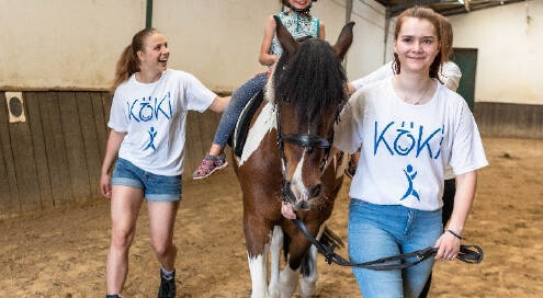 Ein lächelndes Mädchen reitet auf einem Pferd, begleitet von zwei Betreuerinnen in 'KÖKI'-T-Shirts, die das Pferd in einer Reithalle führen.