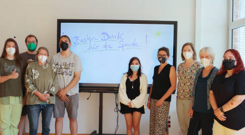 Gruppe aus neun Schülern und Lehrern vor einer digitalen Tafel, alle tragen Mundschutzmasken, auf der Tafel steht „Besten Dank für die Spende!“