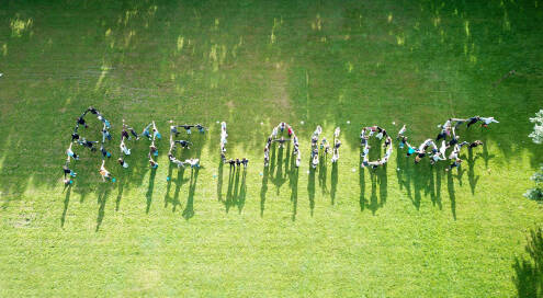 Rasenfläche, Foto von oben aufgenommen, viele Kinder stehen auf dem Rasen, halten sich an den Händen und bilden das Wort „Ameland“. Es ist sonnig, drumherum Bäume.